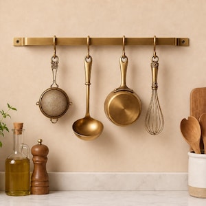 Könnte beinhalten: Eine messingfarbene Küchenleiste an der Wand mit vier hängenden Utensilien: ein rundes Teesieb, eine tiefe Schöpfkelle, ein kleiner Topf und ein Schneebesen. Darunter stehen eine Ölflasche aus Glas, eine Pfeffermühle und ein Keramiktopf mit Löffeln.