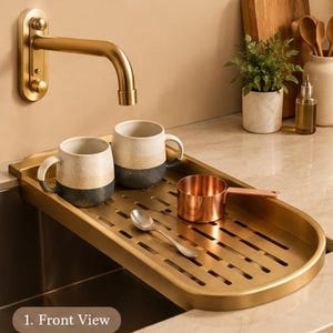 May include: A gold-toned metal sink tray with a slotted surface, featuring two two-toned ceramic mugs, a small copper-coloured measuring cup, and a silver spoon. A wall-mounted gold faucet is positioned above the tray, which displays the text 1. Front View.