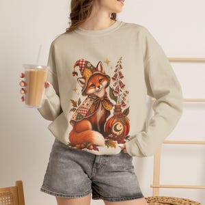 May include: A beige long-sleeve sweatshirt featuring a graphic print of a fox wearing a patchwork hat and cape, standing next to a bottle with a crescent moon symbol and tall flowers. The fox is surrounded by autumn leaves and small stars.