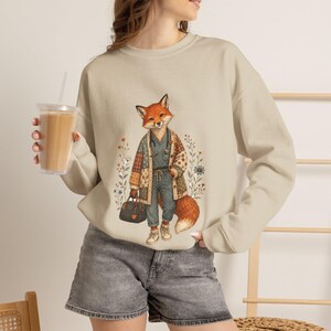 May include: A beige crewneck sweatshirt featuring a graphic illustration of a fox dressed in a patterned cardigan, scrub top, and trousers, holding a dark handbag. The fox is surrounded by small floral accents. The sweatshirt has long sleeves.