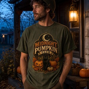 May include: An olive green short-sleeve t-shirt featuring a graphic print with the text "MIDNIGHT PUMPKIN CLUB" in a decorative frame. The design includes a black cat, three orange pumpkins, autumn leaves, and a crescent moon against a starry sky.