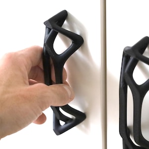 May include: Two matte black cabinet handles with an open, geometric, lattice-like structure. The handles are mounted vertically on white cabinet doors, featuring a complex, interconnected frame design that creates a series of triangular voids.