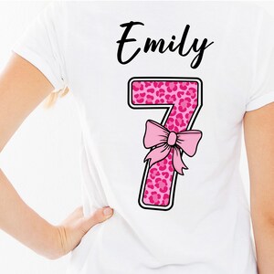 Könnte beinhalten: Ein weißes T-Shirt mit dem Namen Emily in schwarzer Schreibschrift über einer großen Zahl 7. Die Zahl 7 hat ein pinkes Leopardenmuster und ist mit einer hellpinken Schleifengrafik verziert. Das Design ist auf dem Rücken des T-Shirts zentriert.
