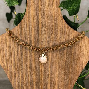 Puede incluir: Un collar tipo gargantilla de macramé marrón que presenta una serie de pequeñas cuentas de madera de color marrón claro tejidas en el cordón. Un colgante circular de concha rosa claro con un engaste de metal dorado cuelga del centro.