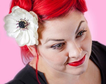 May include: A woman with bright red hair styled in pin curls wears a black dress and a white flower in her hair. She is looking down and smiling.