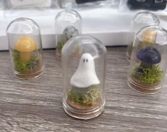 May include: Four glass jars with cork tops contain miniature scenes. One jar has a yellow mushroom, one has a white ghost, one has a brown mushroom with a purple cap, and one has a speckled brown mushroom.