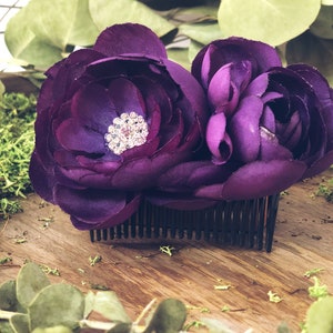 May include: A dark purple floral hair comb featuring two large fabric flowers with a sparkling rhinestone center. The comb is black and set against a wooden surface with greenery.