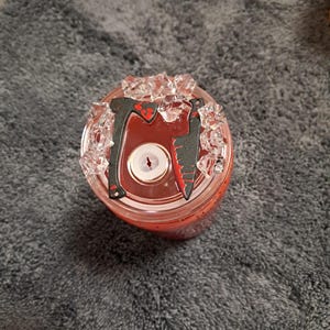 May include: A clear plastic tumbler lid featuring a central straw opening, decorated with miniature black axe and knife charms accented with red paint, surrounded by small, clear, faceted acrylic ice-like pieces on the top surface.