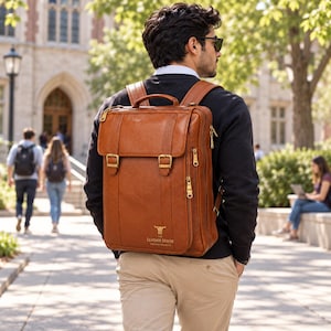 Puede incluir: Una mochila de cuero color canela con asa superior, dos correas con hebilla en la parte delantera y cremalleras doradas. La parte inferior frontal muestra un logotipo dorado con el texto "THE LEATHER HOUSE PREMIUM PRODUCTS". Correas ajustables.