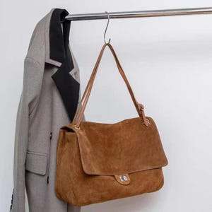 Könnte beinhalten: Eine braune Umhängetasche aus Wildleder mit Umschlagklappe und einem silberfarbenen Metallverschluss. Die Tasche hat einen langen, verstellbaren braunen Riemen und hängt an einem Metallhaken neben einem grauen Mantel mit schwarzem Revers.