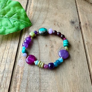 May include: A colorful beaded bracelet with purple, green, blue, and turquoise stones. The bracelet is made with a variety of different shapes and sizes of beads.