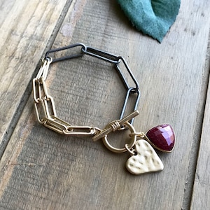 May include: A gold and silver-toned chain bracelet with a toggle clasp. The bracelet features a gold heart charm and a triangular red gemstone charm. The bracelet is displayed on a wooden surface.