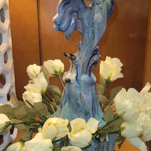 May include: A ceramic sculpture featuring a stylised blue and green seahorse perched atop a fluted, striped base. The base is filled with artificial cream-coloured roses with green leaves, and the entire piece sits inside a dark blue scalloped saucer.