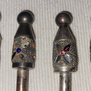 May include: Four metal cocktail picks with spherical tops and textured cylindrical handles. Each handle features a four-petaled floral inlay design with colorful embedded stones in red, blue, pink, and green, set against a silver-toned metal finish.