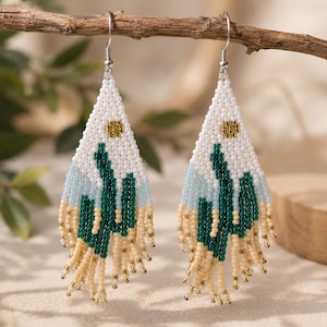 Peut inclure: Une paire de boucles d'oreilles triangulaires en perles représentant un paysage désertique. Le motif comprend un ciel blanc avec un soleil doré, des montagnes bleu clair, des cactus vert foncé et un sol beige avec des franges de perles.