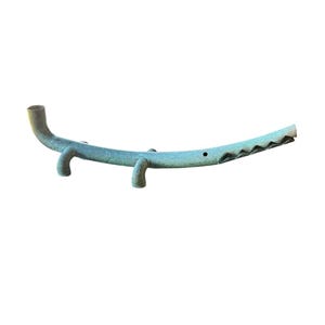May include: A long, slender, teal-coloured ceramic sculpture shaped like a stylised crocodile. The object features a curved body, four short legs, a small circular eye, and an open, jagged mouth. The surface has a matte, weathered texture.