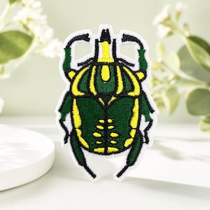 May include: An embroidered patch shaped like a beetle with a dark green body and bright yellow striped markings. The insect design features black outlines and a white border, showcasing detailed stitching throughout the entire beetle figure.