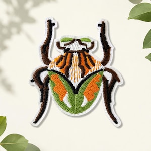 May include: An embroidered patch shaped like a beetle with a white border. The insect features a vibrant color palette of bright orange, lime green, white, and dark brown. The design includes symmetrical patterns on the wings and six dark brown legs.