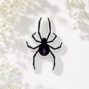 Peut inclure: Un écusson brodé en forme d'araignée avec un corps et des pattes noirs bordés de fil blanc. L'araignée présente une petite marque violette en forme de croix verticale centrée sur son abdomen, créant un contraste net avec le noir.
