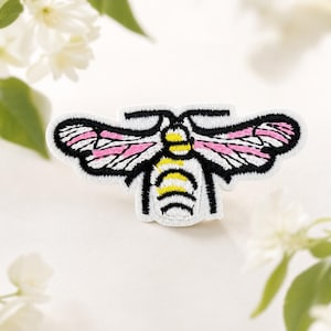 Op de afbeelding: Een geborduurde patch in de vorm van een mot met een wit lichaam, een geel en zwart gestreept achterlijf en vleugels met zwarte omlijningen, witte secties en roze accenten. Het ontwerp is symmetrisch met twee antennes bovenaan.
