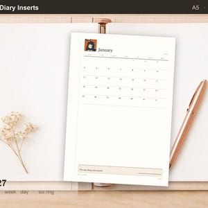 Può includere: Un inserto per agenda datato 2027 in formato A5 con un calendario di gennaio e una piccola illustrazione di un gatto in alto a sinistra. La pagina include una sezione etichettata The one thing this month in basso, su un'agenda aperta.