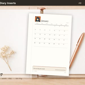 Op de afbeelding: Een A6 gedateerde 2027 agenda-inlegvel met een januari-kalender, een kleine cartoon kat-illustratie linksboven, een sectie voor het doel van de maand en een roségouden pen op een open beige ringband met zes ringen.