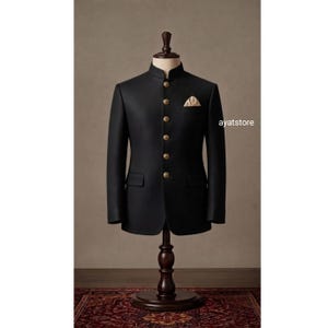 Puede incluir: Una chaqueta bandhgala negra con cuello mao, mangas largas y cierre frontal con cinco botones dorados. La chaqueta incluye dos bolsillos laterales con solapa y un pañuelo de bolsillo beige. El texto ayatstore es visible.