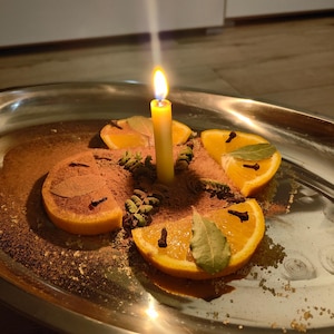 Op de afbeelding: Een brandende gele kaars in het midden van een zilveren ovale schaal, omringd door sinaasappelschijfjes, verspreid bruin kaneelpoeder, gedroogde groene laurierblaadjes en hele kruidnagels in een cirkelvormig patroon op het metalen oppervlak.