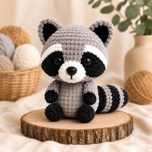 May include: A crocheted raccoon plush toy featuring a grey body, black eye patches, a white muzzle, and a black nose. The raccoon has large black plastic eyes, small rounded ears with black centers, and a striped tail, all crafted from yarn.