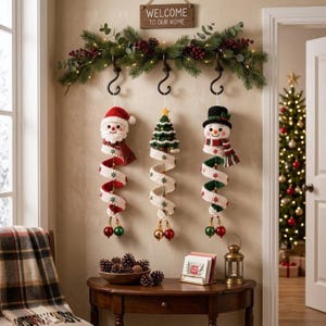 May include: Three crocheted Christmas ornaments hanging from black hooks on a green garland with red berries and lights. The ornaments feature a Santa Claus, a Christmas tree, and a snowman, each with a long, spiraling white tail with green snowflakes.