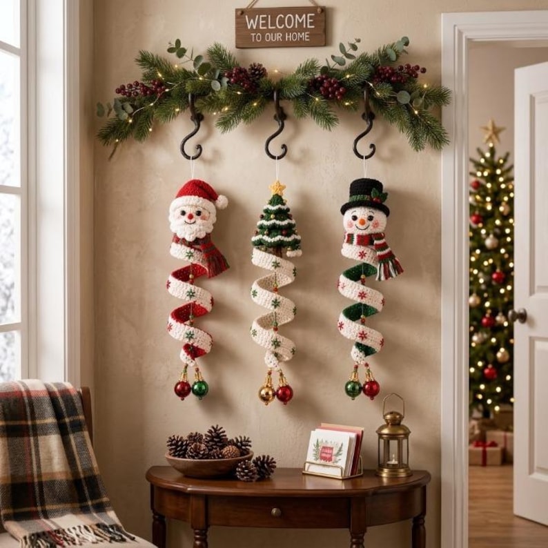May include: Three crocheted Christmas ornaments hanging from black hooks on a green garland with red berries and lights. The ornaments feature a Santa Claus, a Christmas tree, and a snowman, each with a long, spiraling white tail with green snowflakes.