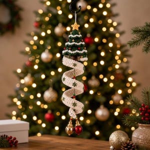 May include: A crocheted Christmas tree ornament featuring a dark green tree with white trim and a yellow star topper. A long, spiraling cream-colored ribbon with embroidered green snowflakes hangs below, accented by small beads and two shiny round bells.