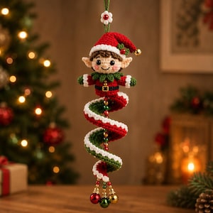 Puede incluir: Un adorno navideño de elfo tejido a ganchillo con un gorro rojo y blanco, un cuello verde y un cinturón negro con hebilla dorada. El elfo está unido a una larga cola en espiral tejida en rojo y verde, decorada con pequeñas campanas rojas, verdes y doradas.