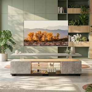 May include: A rectangular television stand with a light gray concrete-textured finish, featuring a floating top shelf supported by clear pillars, an open middle glass shelf, and a lower storage area containing small decorative objects and a radio.