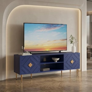 May include: A navy blue media console featuring two side cabinets with geometric carved patterns and gold diamond-shaped pulls. The unit has a central open shelf, four slender gold legs, and supports a flat-screen television displaying a sunset.