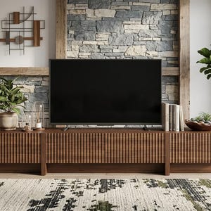 May include: A long, rectangular media console crafted from dark brown wood featuring a vertically fluted, ribbed texture across its front panels. The console is supported by a low base and provides a flat surface for a television and decor.
