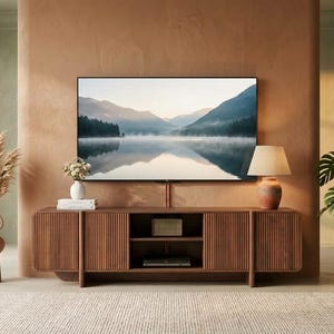 May include: A long, rectangular wooden media console with vertical slatted cabinet doors, featuring two open central shelves. The console is positioned beneath a wall-mounted television displaying a landscape of mountains and a lake.
