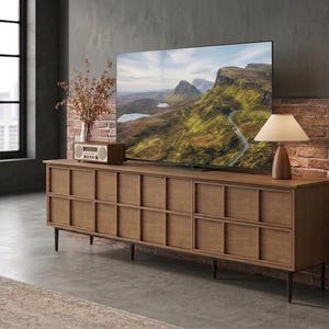 May include: A long wooden media console with a grid-patterned front, supported by thin black legs. The top surface holds a large flat-screen television displaying a mountain landscape, a small wooden radio, and a lamp with a white shade.