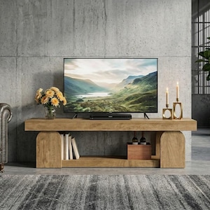 May include: A light brown wooden media console with two arched side supports and a lower shelf. The console holds a flat-screen television displaying a mountain landscape, a glass vase with yellow flowers, and two gold candle holders with candles.