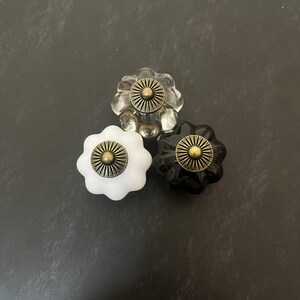 Op de afbeelding: Drie bloemvormige kastknoppen gerangschikt in een driehoek. Eén knop is van helder glas, één is effen wit en één is effen zwart. Elke knop is voorzien van een ronde, antiek messingkleurige metalen middenplaat met een zonnestraalpatroon.