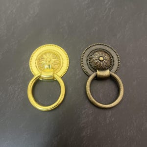 Könnte beinhalten: Zwei kreisförmige Möbelgriffe mit schwingendem Ring. Ein Griff hat ein glänzendes Gold-Finish, während der andere eine gealterte, dunkle Bronzepatina aufweist. Beide Stücke zeigen ein zentrales Blumenmotiv und einen perlenartigen Rand.