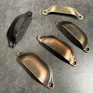 Può includere: Una collezione di cinque maniglie a conca per cassetti in metallo con varie finiture, tra cui nero opaco, ottone spazzolato, bronzo antico e rame. Ogni maniglia presenta un design curvo semicircolare con due fori per le viti alle estremità.