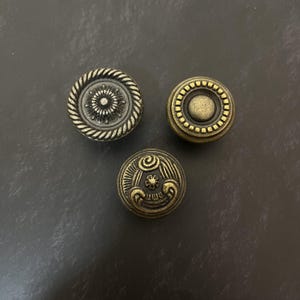 Peut inclure: Trois boutons de placard circulaires en métal avec une finition en laiton antique. Le bouton en haut à gauche présente une bordure en forme de corde et un motif floral central. Le bouton en haut à droite a un bord perlé et un centre lisse.