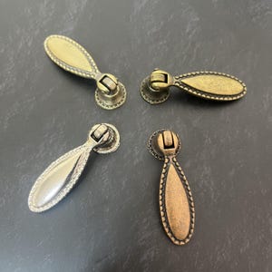 Könnte beinhalten: Vier hängende Möbelgriffe aus Metall, jeder mit einem tropfenförmigen Griff und einem dekorativen Perlenrand. Zwei Griffe haben ein mattes Messing-Finish, einer ein helles silberfarbenes Finish und einer ein dunkles kupferfarbenes Finish.