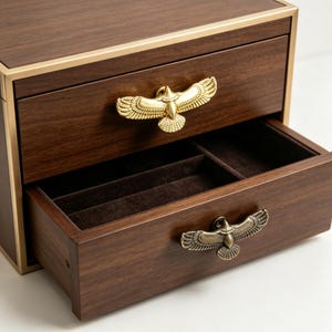 May include: A dark wood jewelry box featuring two drawers with gold-toned eagle-shaped metal handles. The top drawer is closed, while the bottom drawer is open, revealing a dark brown velvet-lined interior divided into two separate storage compartments.