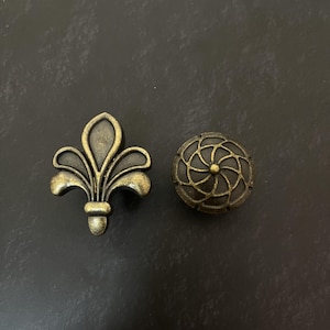 May include: Two metal cabinet knobs with an antique brass finish. One knob is shaped like a fleur-de-lis with a dark recessed centre. The other knob is circular with a raised, swirling, interconnected line pattern and a dark recessed background.