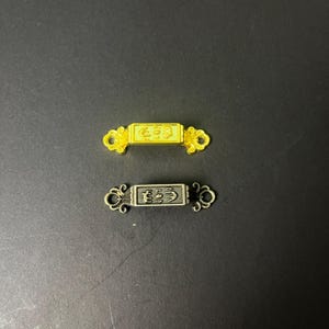 May include: Two rectangular metal connector charms, one in a bright gold tone and one in a dark gunmetal tone. Each piece features a central rectangular bar with embossed decorative symbols and ornate, swirling filigree loops on both ends.