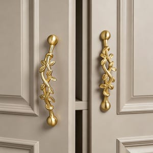 Può includere: Due maniglie per armadietto in metallo color oro con un motivo a vite e foglie. Ogni maniglia presenta un design a stelo centrale intrecciato decorato con piccole foglie in rilievo ed estremità arrotondate, montate verticalmente su ante di armadietto grigio chiaro.