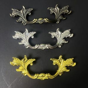 May include: Three ornate metal drawer pulls arranged vertically. The top pull is dark bronze, the middle pull is polished silver, and the bottom pull is bright gold. Each handle features intricate, symmetrical scrolling leaf patterns.