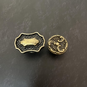 May include: Two metal buttons with an antique brass finish. One button is rectangular with a scalloped edge and a textured grid pattern surrounding a smooth centre. The second button is circular with a raised, symmetrical floral-like design.